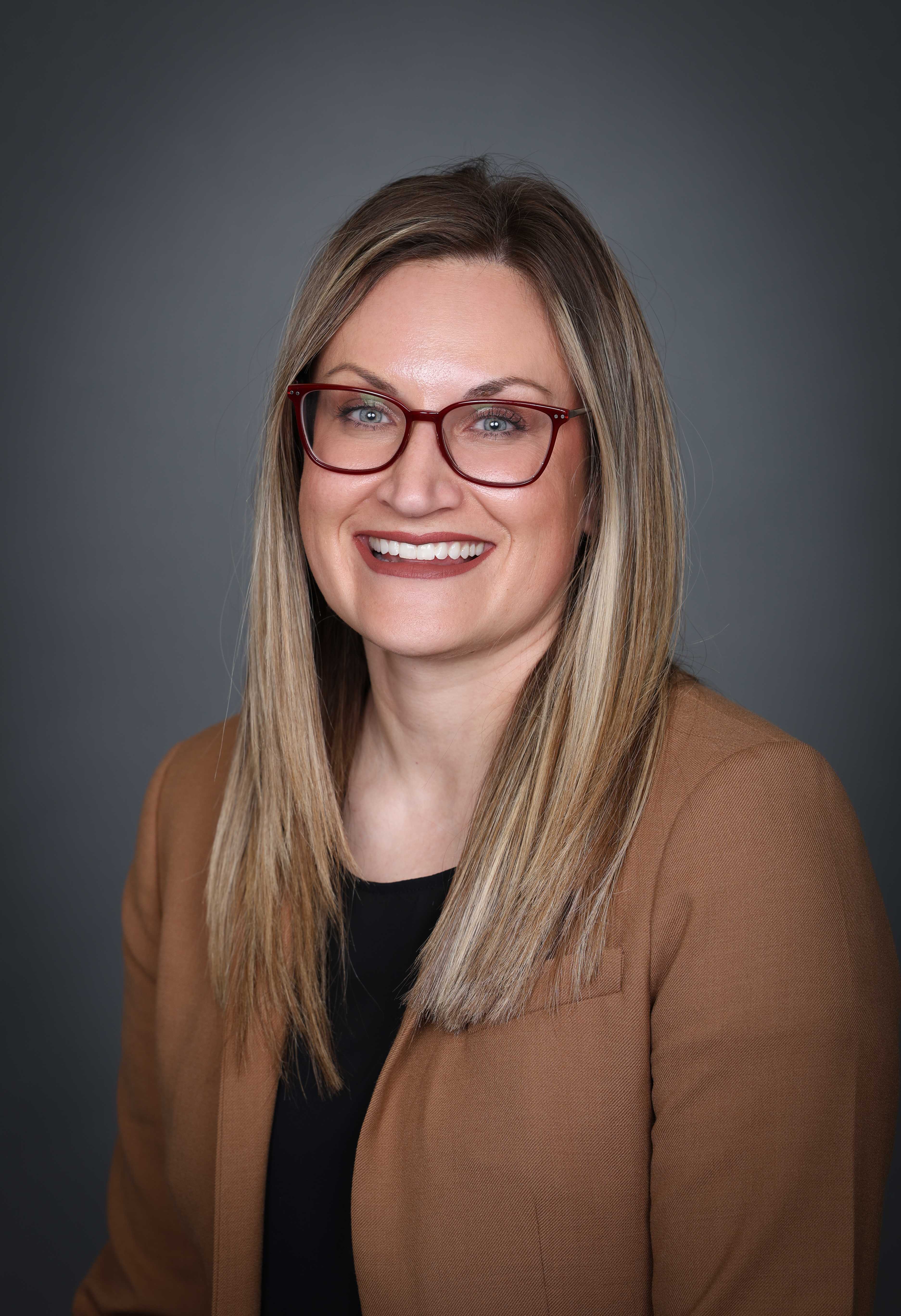 photo of Madi Koenig with long blondish brown hair a black shirt, brown jacket and glasses